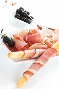 Delicious prosciutto ham background with black olives, fresh herbs and grissini breadsticks.