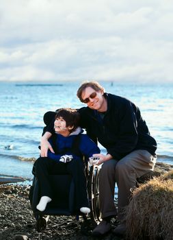 Father sitting with disabled son in wheelchair by lake shore