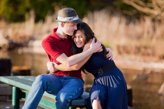 Young Caucasian man hugging his biracial girlfriend, sitting together on dock over lake