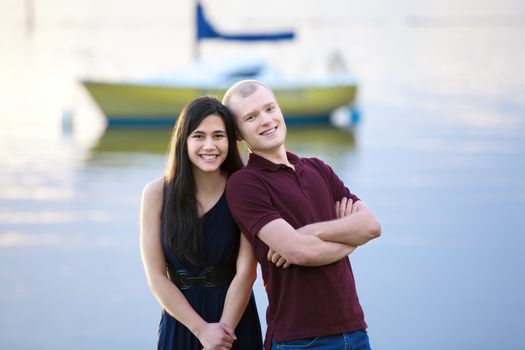 Happy young interracial couple standing together by lake, colorful boat in background