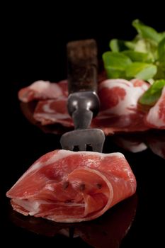 Parma ham prosciutto on old fork, ham slices with green salad in background isolated on black. Culinary meat eating.