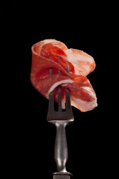 Delicious parma ham slice on old silver fork isolated on black background. Culinary traditional italian parma ham concept. Prosciutto.