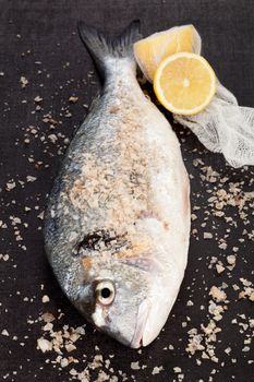 Fresh fish, sea salt crystal and cut lemon isolated on textured black background. Luxurious mediterranean seafood concept.