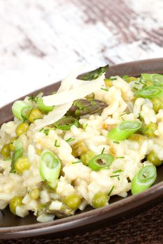 Delicious risotto with peas, asparagus, onion and parmesan cheese on brown plate on old white wooden background. Culinary vegetarian food.