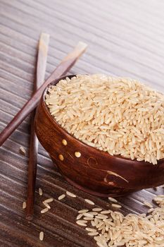 Natural brown rice background. Raw uncooked rice in wooden bowl with chopsticks on brown background. Culinary rice eating.