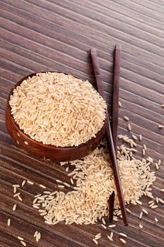 Brown natural rice in oriental wooden round bowl with wooden chopsticks on brown background. Culinary healthy eating background.