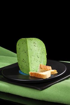 Green pesto gouda cheese piece on black plate on black background. Culinary cheese eating.