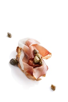 Delicious canape with prosciutto, capers and dry tomatoes isolated over white. Culinary eating.