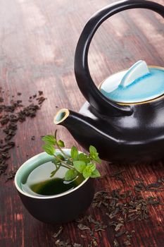 Traditional tea background. Tea in tea bowl with fresh mint herb, ceramic tea pot, dry tea crop on brown wooden background. Asian tea drinking ceremony.