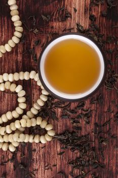 Traditional asian tea ritual. Dry green tea crop, hot tea in cup and buddhist necklace on brown wooden background.  