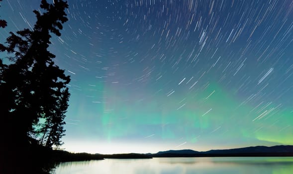 Astrophotography star trails on midsummer night sky with Aurora borealis or Northern Lights over calm water surface of Lake Laberge, Yukon Territory, Canada