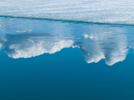 Global warming concept of receding ice sheet and white clouds reflected on calm blue arctic water