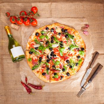 Delicious tasty pizza, fresh tomatoes, garlic, bottle of white wine and wooden cutlery on brown background, top view. Rustic country style italian eating concpept.