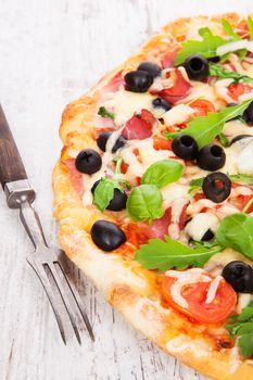 Delicious colorful pizza with tomatoes, melted cheese, black olives, onions, ham and fresh herbs on white wooden background. Culinary mediterranean pizza eating.