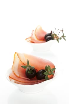 Prosciutto slices with black olives and fresh herbs in white bowl isolated on white background. Luxurious culinary canape.