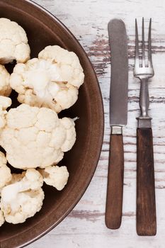 Fresh raw cauliflower on dark brown plate with wooden cutlery on white wooden textured background, top view. Fresh raw vegan and vegetarian eating concept. 