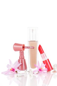 Luxurious makeup and cosmetic still life. Nail polish, red lipstick and beige foundation with pink and white flowers isolated on white background with reflection. Glamour beauty concept.