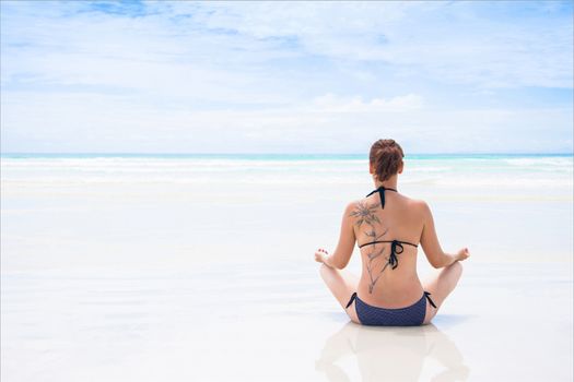 Rear view of beautiful young woman with tattoo doing yoga on beach. Wellbeing concept.