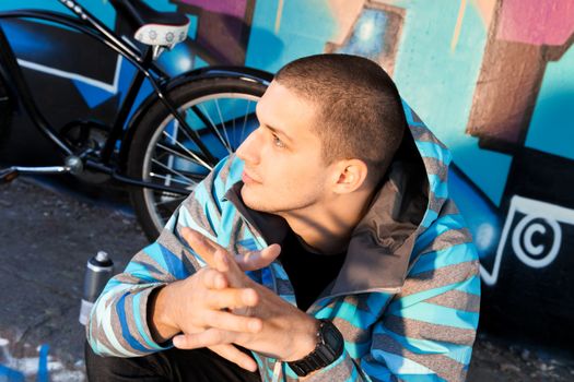 Young male graffiti artist sitting in front of finished piece on wall with city cruiser bike leaning next to him. Urban culture.