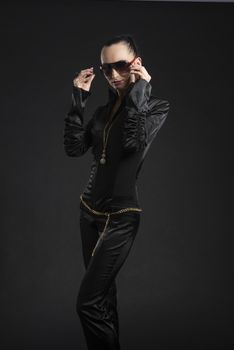 Portrait of beautiful Caucasian woman putting on sunglasses and wearing black bodysuit against black background