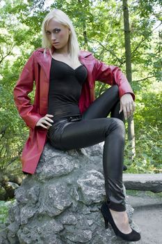 blond sexy woman outside in park sitting on a rock with a red skin coat