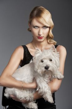 young elegant blond woman wearing black dress with an old fashion hairtyle and necklace jewellery, she looks down at the left and touches her hair with right hand, she is in front of the camera, looks in to the lens and has a dog in her arms.