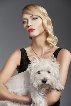young elegant blond woman wearing black dress with an old fashion hairtyle and necklace jewellery, she looks in to the lens with actractive eyes and takes the dog in her arms
