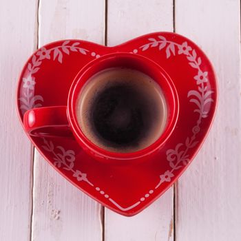A red cup of coffee with heart shaped plate, over white wood