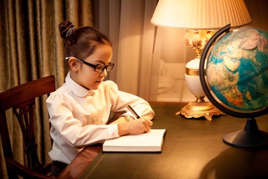 Little girl writing in notebook at desk with lamp and globe