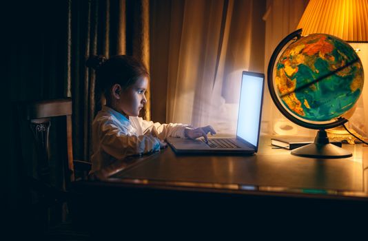Portrait of little girl doing homework on laptop at evening
