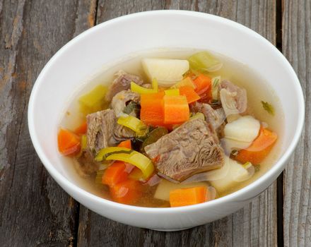 Rustic Beef Soup with Potato, Carrot, Leek and Greens in White Bowl isolated in Wooden background