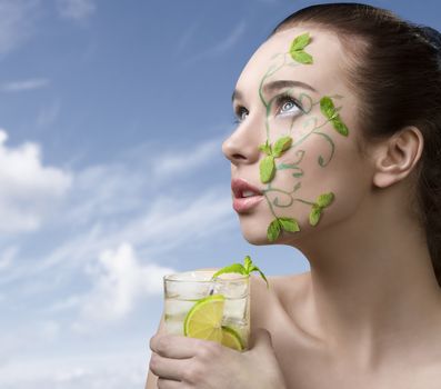 pretty girl with brown hair having creative green make-up with some mint leaves, naked shoulder and mojito cocktail in the hand