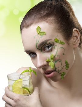 beauty portrait of cute brunette girl with creative green make-up and some mint leaves on the face, taking a cocktail mojito in hand