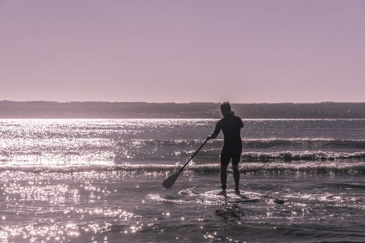 Young unrecognizable man paddle surfs the waves in Majorca, Balearic islands, Spain. Sunny violet filter treatment.
