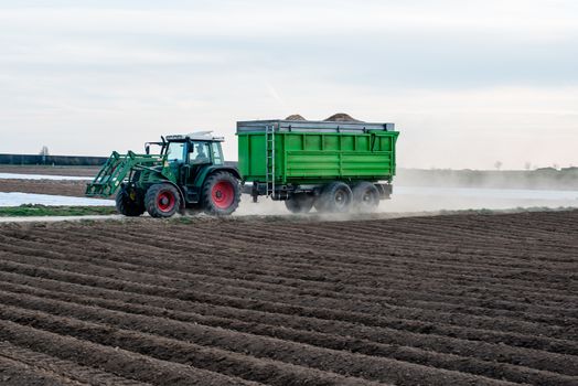 Modern tractor with trailer driving along agricultural field and raising dust due to dry weather conditions in early spring