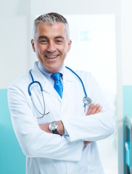 Friendly doctor portrait with arms crossed and medical equipment in teh background.