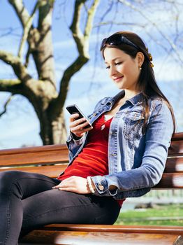 Photo of a beautiful young woman using a smartphone while sitting in a park on a bench.