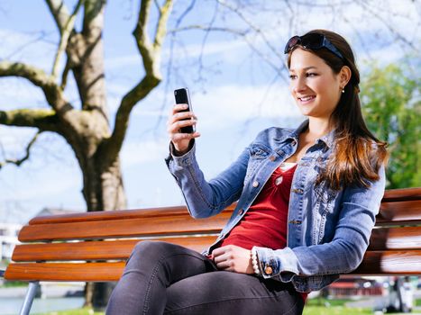 Photo of a beautiful young woman using a smartphone while sitting on a park bench.
