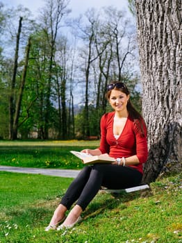 Photo of a beautiful young woman reading a book sitting against a tree in early spring.
