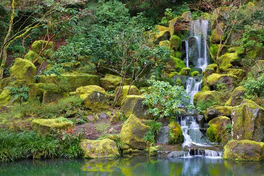 Asian Garden Waterfall flowing into a still pond surrounded by moss covered rocks, trees, and other foliage