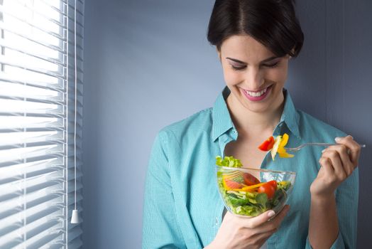 Woman smiling and eating salad in front of a window.