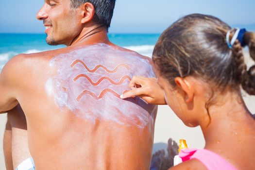 Adorable girl at tropical beach applying sunblock cream on a father's back.