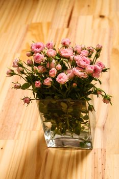 Bunch of small pink Roses in a glass vase over a wooden table.
