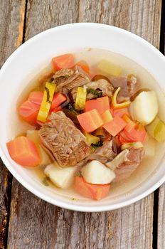 Rustic Beef Soup with Potato, Carrot, Leek and Greens in White Bowl closeup on Wooden background. Top View