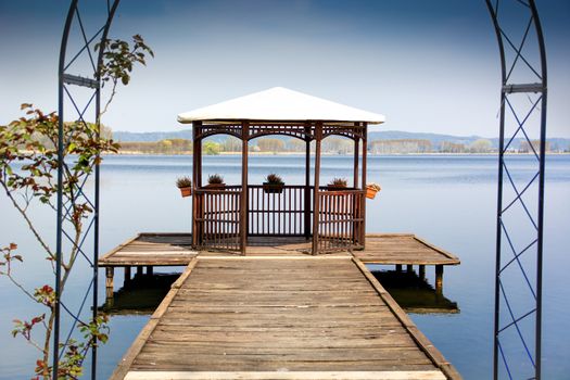 Wooden gazebo on dock over peaceful lake, symmetric shot. Daytime