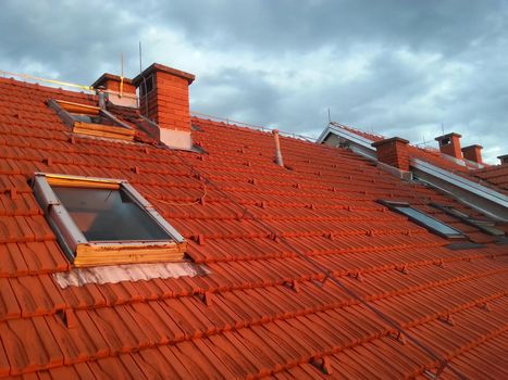 Tiled roof with windows and chimneys and stormy sky in background.