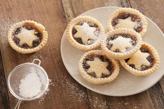 Preparing a plate of delicious Christmas mince pies decorated with pastry stars and sprinkling them with icing sugar from a sieve in a country kitchen on a wooden counter, high angle view