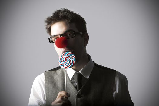 Funny portrait of young stylish man with clown nose eating lollipop against grey background