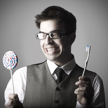 Happy smart young man with a lollipop in one hand and a toothbrush in the other against grey background