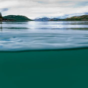Half underwater half over, over-under split shot of huge Lake Laberge, Yukon Territory, Canada, clear blue freshwater and distant shore boreal forest taiga landscape